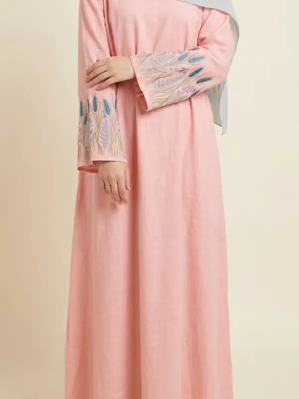 Front view of a Muslim hijabi model wearing a pink linen-viscose maxi dress featuring intricate blue and purple wheat and floral embroidery on the sleeve cuffs.