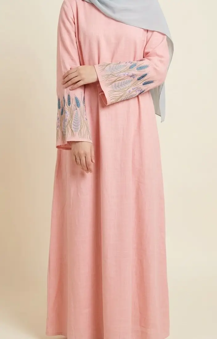 Front view of a Muslim hijabi model wearing a pink linen-viscose maxi dress featuring intricate blue and purple wheat and floral embroidery on the sleeve cuffs.